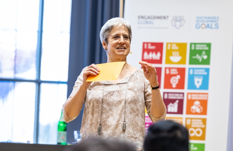 Programme host, Susanne Luithlen. In the background, a banner with the symbols of the 17 Sustainable Development Goals (SDGs).